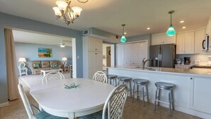 Une cuisine moderne avec des meubles blancs, un îlot central doté de tabourets, un lustre et un coin repas avec une table blanche et des chaises en osier.