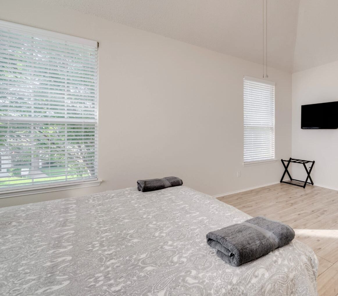 Een slaapkamer met een bed, twee ramen, een televisie en een kleine tafel.