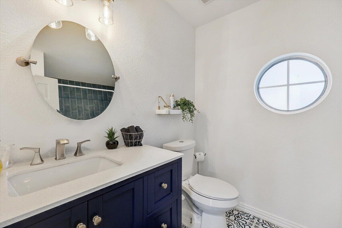 A bathroom with a white sink, dark blue vanity, a round mirror, a toilet, and a small window.