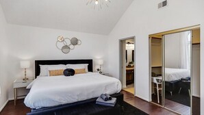 A modern hotel room with a large bed, a desk, a chair, a mirror, and a ceiling light fixture. - Glamorous 4BR 2B Home With Pool, Game Room & Fire Pit (Mesquite)