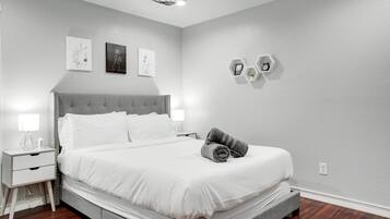Une chambre moderne avec un lit recouvert d’un tissu gris, une literie blanche, une tête de lit grise, des tables de chevet avec des lampes et une étagère hexagonale fixée au mur, sur laquelle se trouve une horloge.