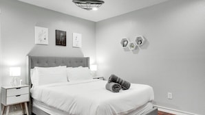 Une chambre moderne avec un lit recouvert d’un tissu gris, une literie blanche, une tête de lit grise, des tables de chevet avec des lampes et une étagère hexagonale fixée au mur, sur laquelle se trouve une horloge.