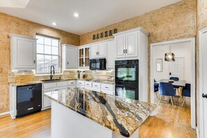 A modern kitchen with white cabinetry, a granite countertop, and a dining area with blue chairs. - Impressive 5BR 3B Home With Pool, Game Room & Cinema Room (Irving)