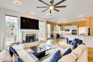 A spacious living room with a fireplace, a large flat-screen TV, and a modern ceiling fan. - Impressive 5BR 3B Home With Pool, Game Room & Cinema Room (Irving)