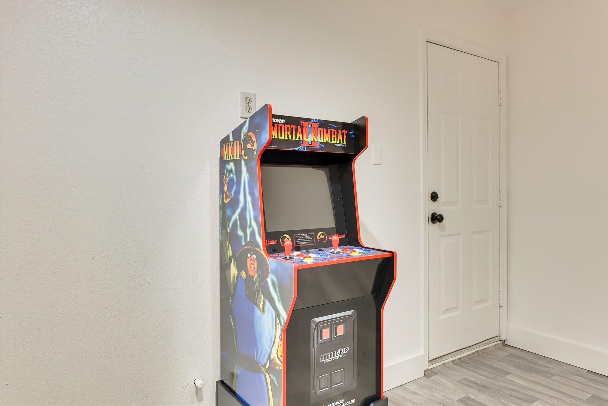 Una máquina de videojuegos retro con el rótulo 'Mortal Kombat' en un pasillo con una puerta blanca y suelo de tonos claros.