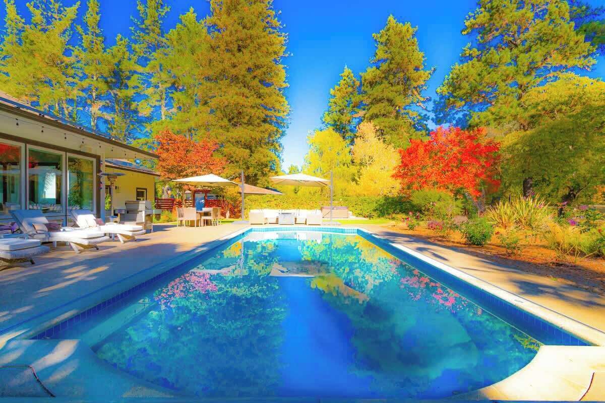 A swimming pool with lounge chairs and umbrellas, surrounded by trees and a building with large windows.