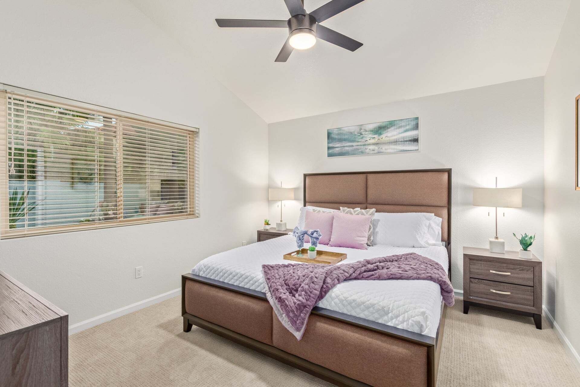 A bedroom with a bed, a ceiling fan, a window with blinds, a bedside table with a lamp, and a wall-mounted artwork.
