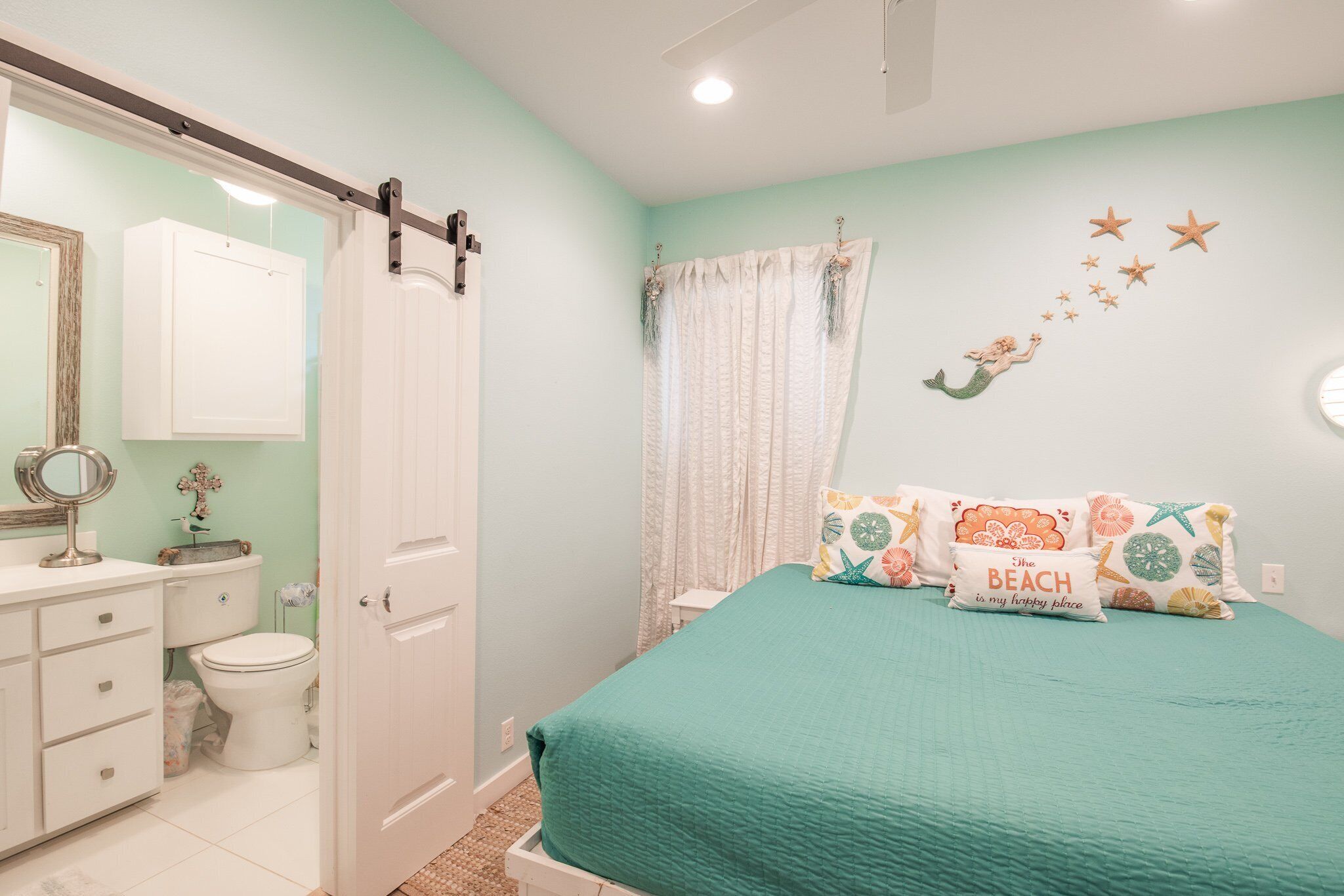 A bedroom with a teal bedspread, a white door, a bathroom with a toilet and sink, and a wall with starfish and mermaid decorations.