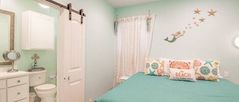 A bedroom with a teal bedspread, a white door, a bathroom with a toilet and sink, and a wall with starfish and mermaid decorations.
