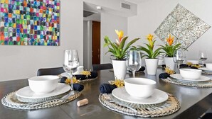 Een eettafel gedekt met witte borden, kommen en glazen, omringd door geweven tafelmatten en potplanten.