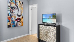 Een gang met een groot abstract schilderij, een televisie op een dressoir en een deur naar een andere kamer.