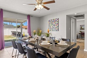 Un espace repas comprenant une longue table, des chaises, un ventilateur de plafond et une vue sur une terrasse extérieure, accessible par des portes vitrées.