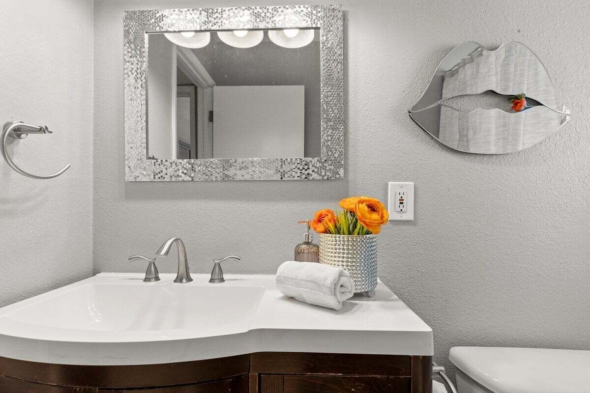 A bathroom with a white sink, two faucets, a mirror with a textured frame, a vase with orange flowers, and a towel.