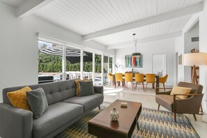 Ein modernes Wohnzimmer mit einem grauen Sofa, einem braunen Sessel und einem Couchtisch. Im Hintergrund ist eine Essecke mit gelben Stühlen und einem Tisch zu sehen.