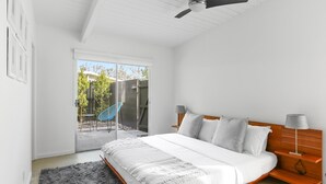 Camera da letto con un letto, una ventola a soffitto, un comodino e vista su una zona esterna con una sedia e piante, tramite una porta scorrevole.