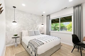 Une chambre moderne avec un grand lit, deux lampes suspendues, une vue sur la verdure et un fauteuil noir.