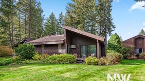 A wooden cabin with a sloped roof, a covered porch with chairs, and a well-maintained lawn. - Twin Lakes Village Home Golf Retreat Community Pool Corner Lot and Single Level Living (Rathdrum)