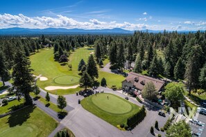 An aerial view of a golf course with a clubhouse, surrounded by dense forest and mountains in the background. - Twin Lakes Village 3-story Townhouse Golf Views Steam Shower Wet Bar Game Room Pool (Rathdrum)