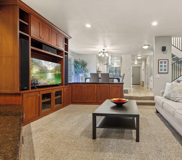 A living room with a TV, wooden cabinets, a sofa, and a staircase. - Twin Lakes Village 3-story Townhouse Golf Views Steam Shower Wet Bar Game Room Pool (Rathdrum)