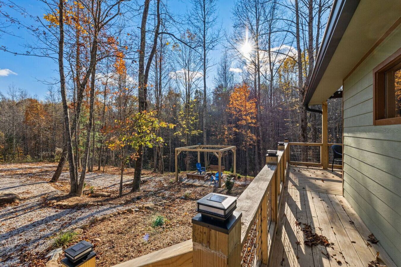 Una terraza de madera con una pérgola y zona de estar, rodeada por un bosque con follaje otoñal.