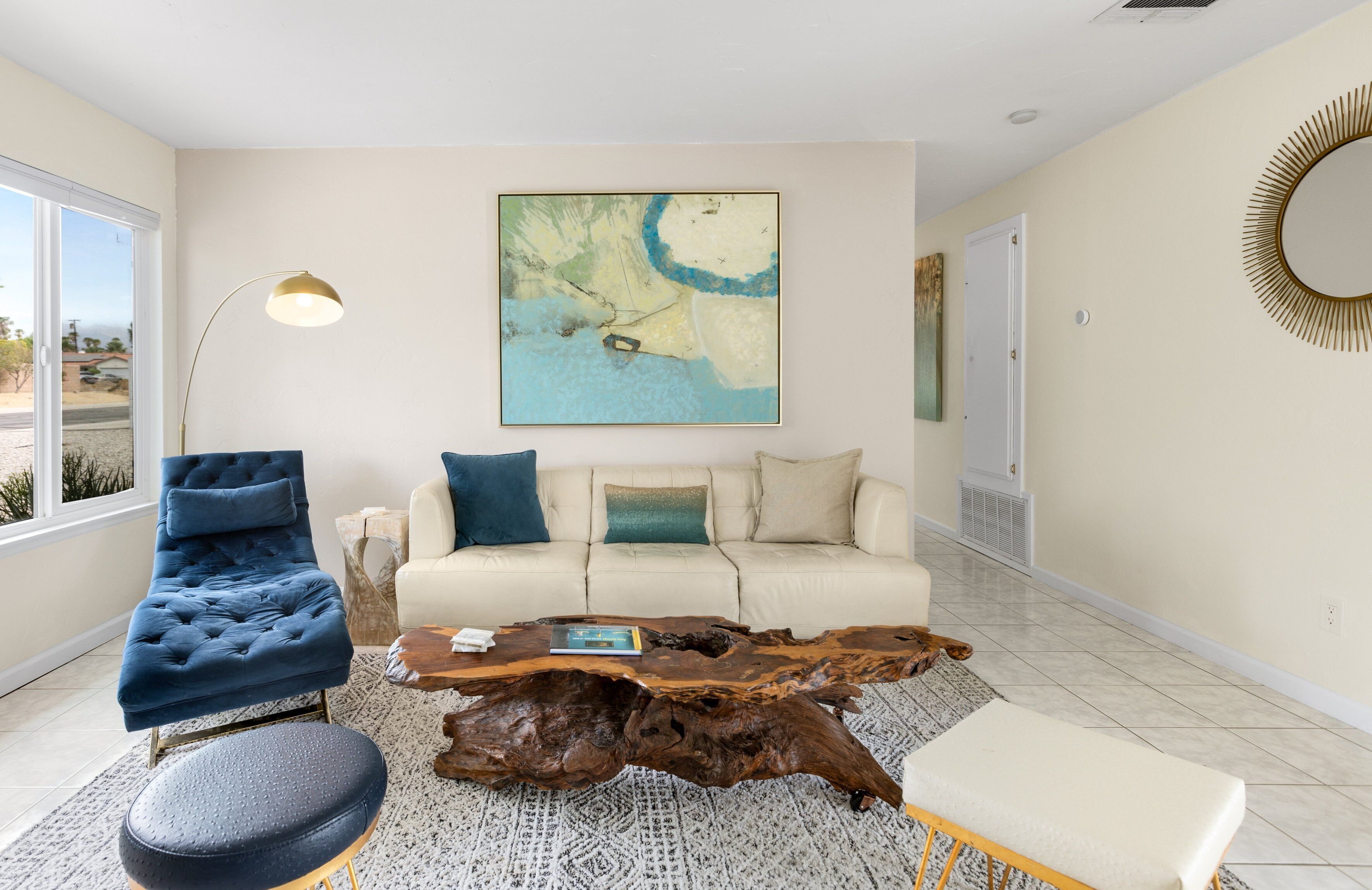 En moderne stue med en blå velourstol, en beige sofa med blå og grønne puder, et træsofabord og et stort abstrakt maleri på væggen.