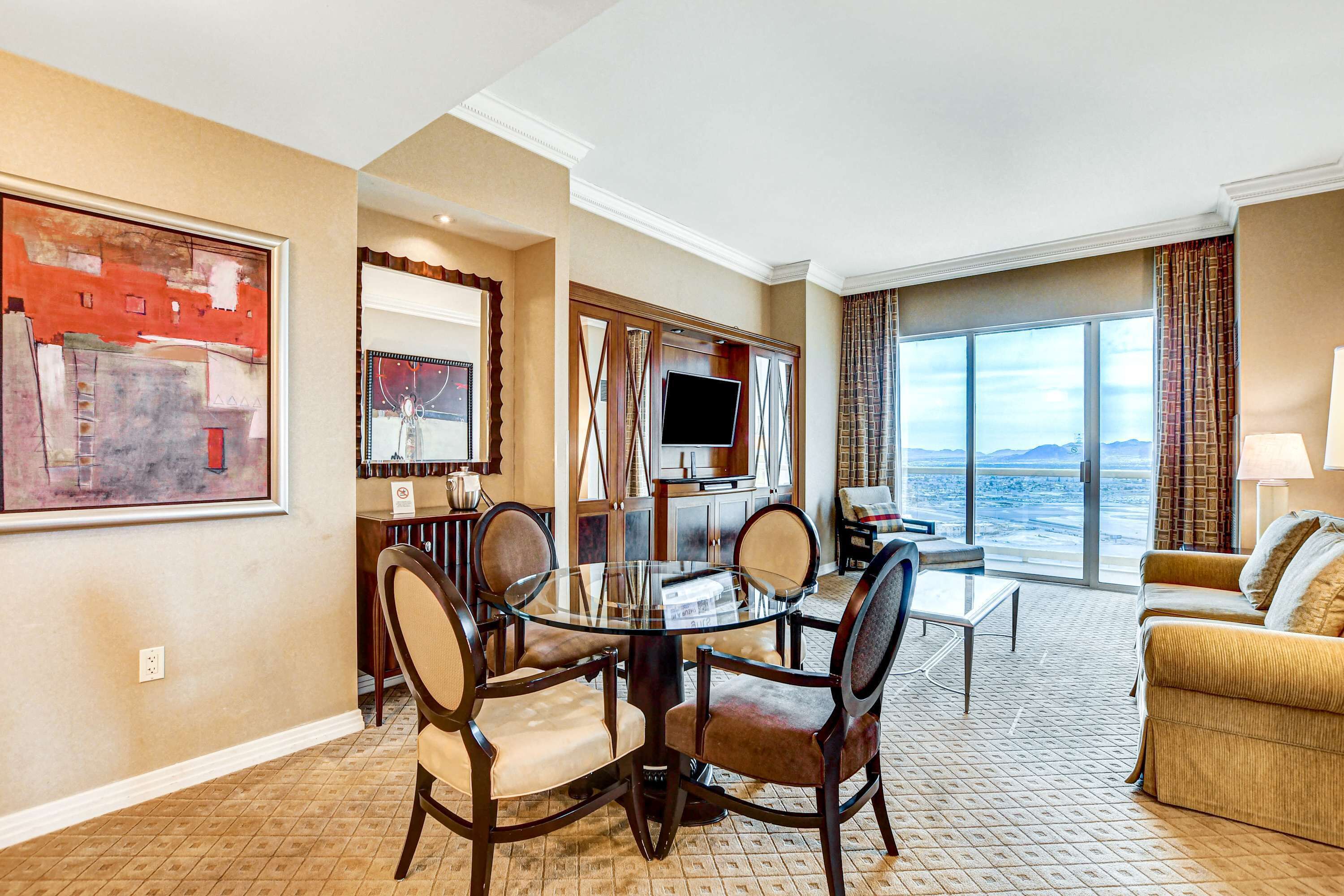 A hotel room with a dining area, a TV, a sofa, and a large window with a view.