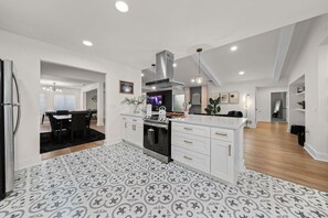 A modern kitchen with white cabinetry, a central island, and a black oven. There is a dining area with a black rug and a TV mounted on the wall. - Spacious 6 Bed House Airport Pet Friendly AFB (San Antonio)