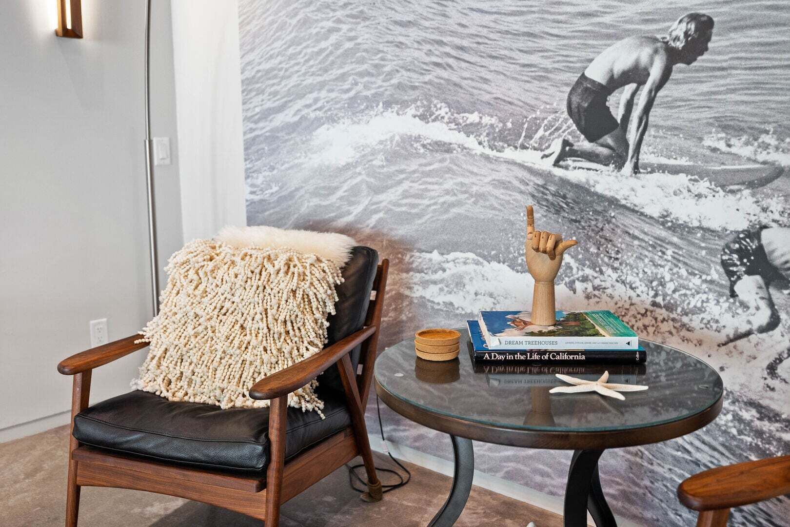 Ein Zimmer mit einem schwarzen Ledersessel, einem Glastisch mit Büchern und einer Holzskulptur einer Hand, sowie einem großen Wandgemälde eines Surfers.