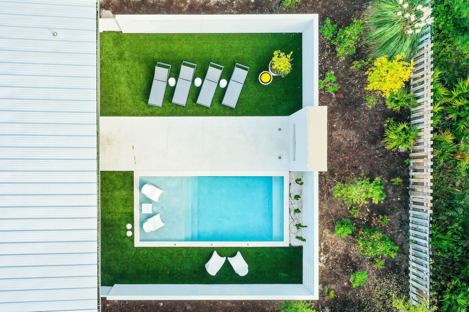 En rektangulär swimmingpool med en poolterrass, omgiven av ett trästaket och frodig grönska.