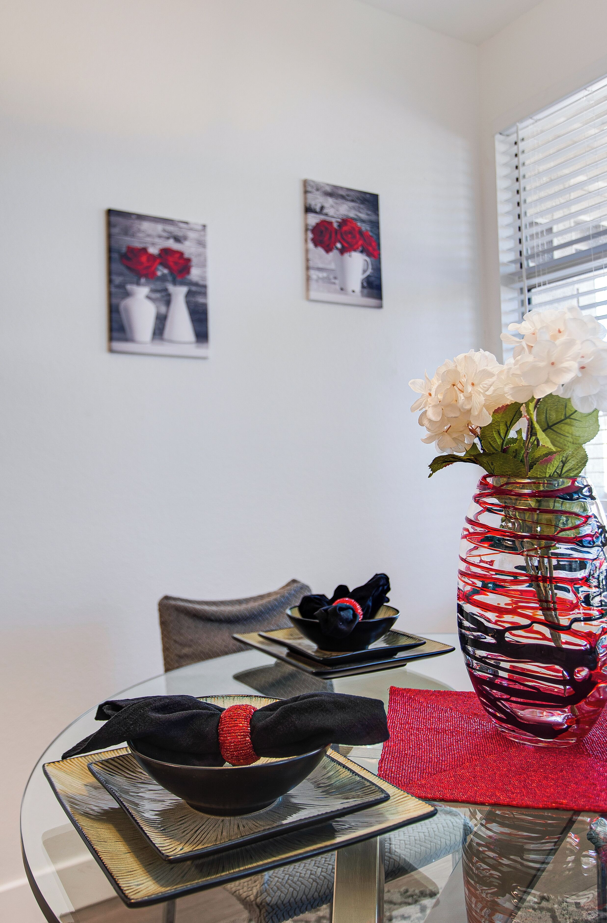 Una mesa de comedor con un florero de vidrio que contiene flores blancas, un cuenco negro y una servilleta roja. Detrás de la mesa hay dos cuadros en la pared.
