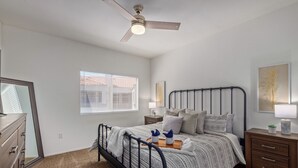 Un dormitorio con cama, mesitas de noche, cómoda, ventilador de techo y ventana con persianas.