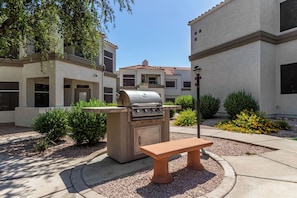 An outdoor barbecue area with a grill and a bench, surrounded by a landscaped garden and a building with multiple windows.