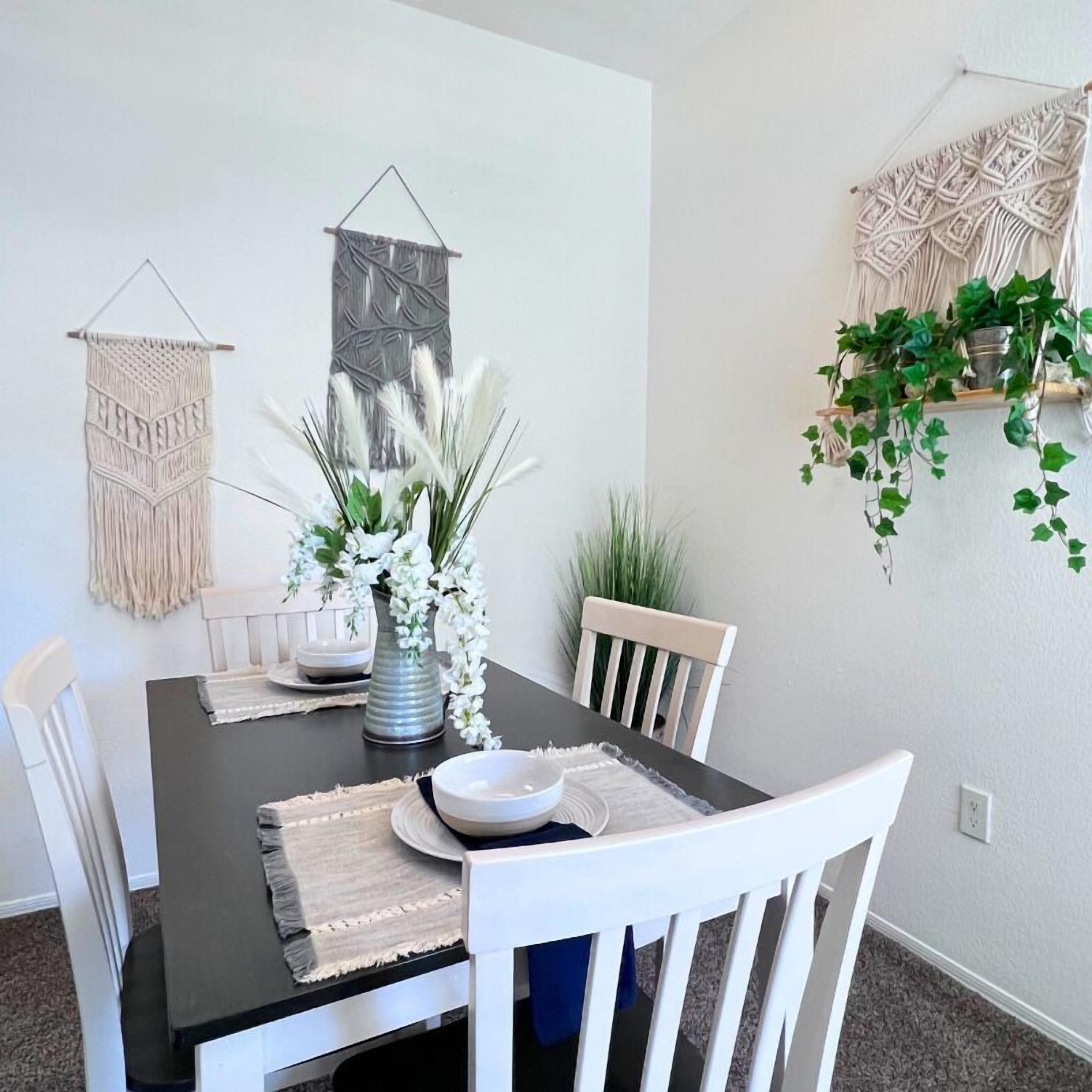 A dining area with a black table, white chairs, a vase with white flowers, and macramé wall hangings.