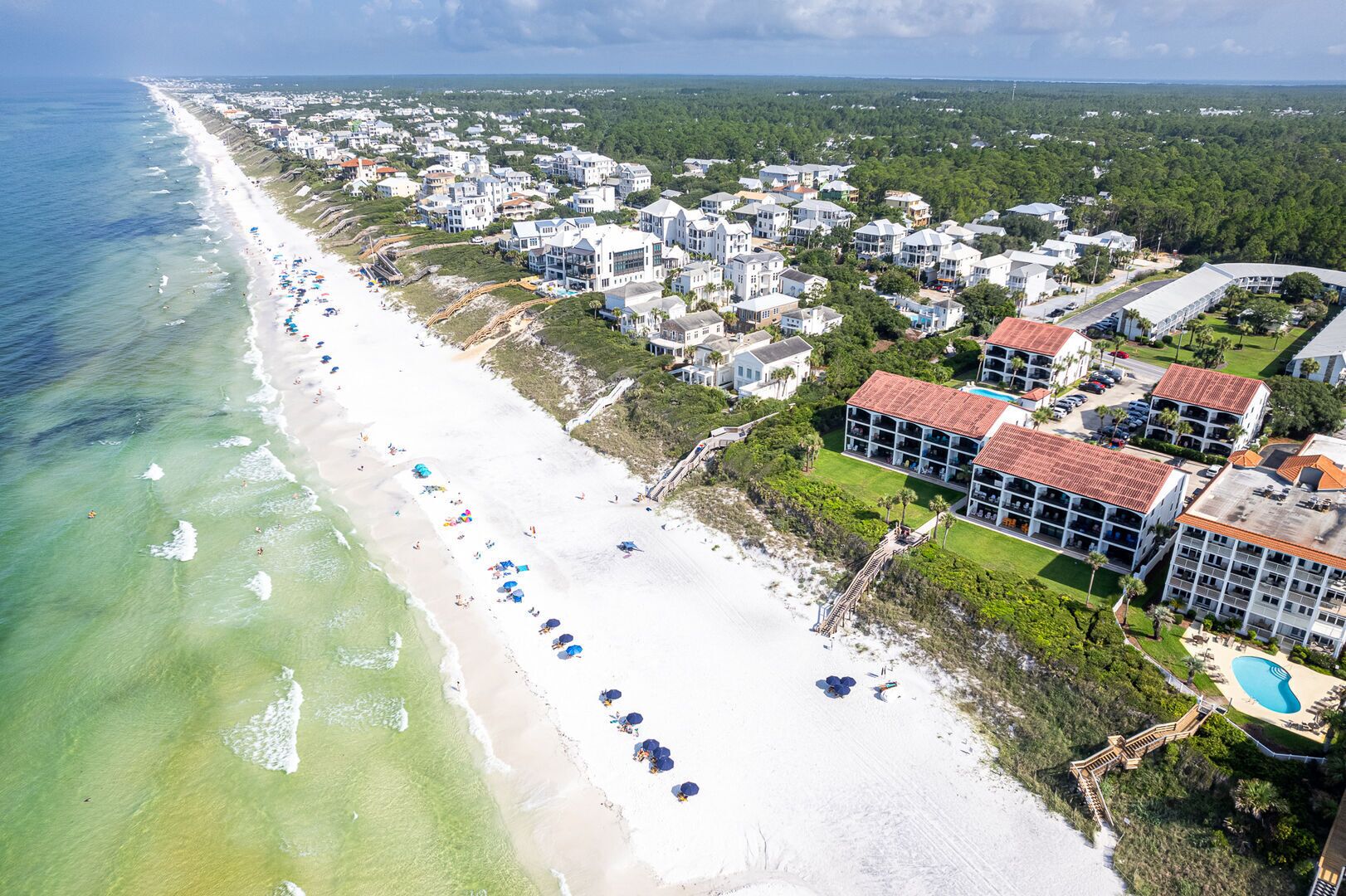 An aerial view of a coastal area with a long sandy beach, a row of beachside buildings, and a large resort complex with multiple buildings and a swimming pool.