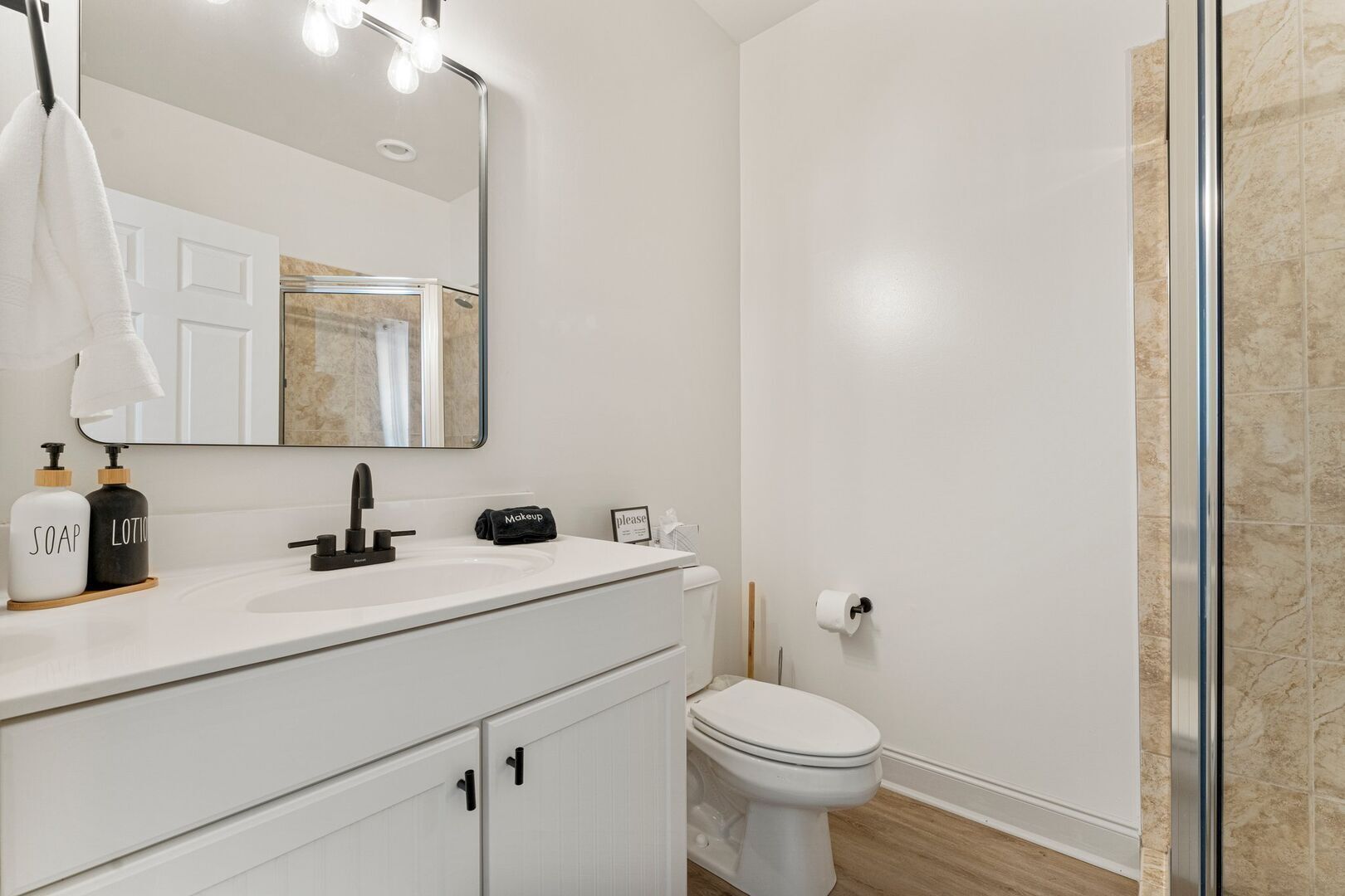 A modern bathroom with a white sink, mirror, and toilet. There are two soap dispensers and a makeup bag on the counter.