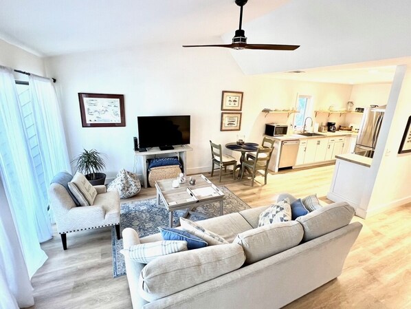 A modern living room with a sofa, coffee table, and TV. There is a dining area with chairs and a table. The kitchen is visible in the background. - Tranquil Oasis Golf Course Views Near Beach 1809 (Seabrook Island)