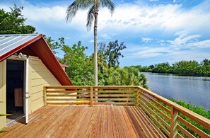 Une terrasse en bois dotée d’une balustrade, qui offre une vue sur une étendue d’eau, le tout entouré d’une végétation luxuriante et sous un ciel dégagé.