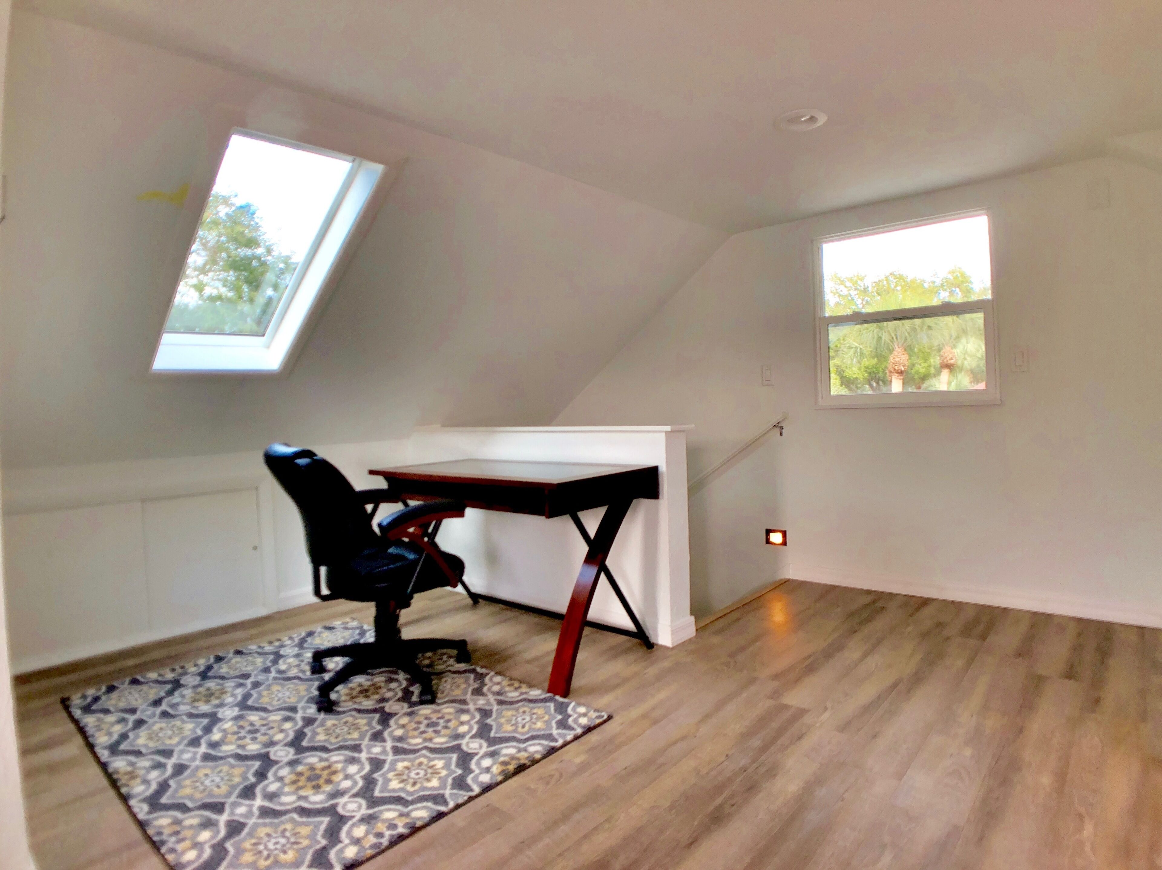 Une petite pièce avec un plafond en pente, comprenant un bureau et une chaise, deux fenêtres et un tapis à motifs.