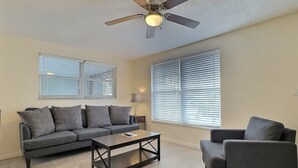 A living room with a grey sofa, a coffee table, and a chair. There is a ceiling fan and a window with blinds. - One Ocean - A (Siesta Key)