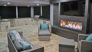 An outdoor patio with a stone floor, wicker furniture, a built-in grill, and a large TV mounted on the wall. - The Treehouse (Siesta Key)