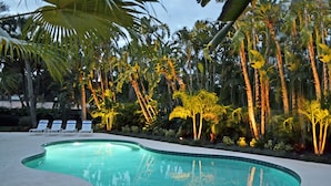 A swimming pool with a curved edge, surrounded by palm trees and other tropical vegetation. - Shell Beach Home (Siesta Key)