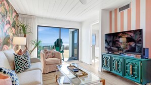 A modern living room with a sofa, armchair, and a glass coffee table. There is a television on a teal cabinet and a large window offering a view of the outdoors. - Seagroovy (Santa Rosa Beach)