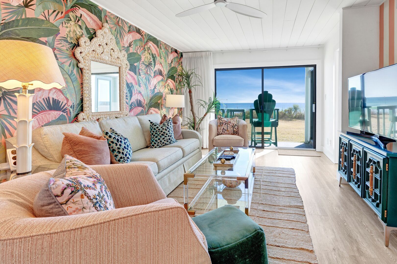 A living room with a sofa, coffee table, and a television. There is a large window with a view of the beach.