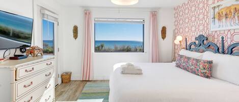 A bedroom with a large bed, a dresser, a television, and a window with a view of the ocean.