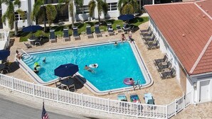 Une piscine rectangulaire entourée de chaises longues et de parasols, avec une clôture blanche et un drapeau américain.