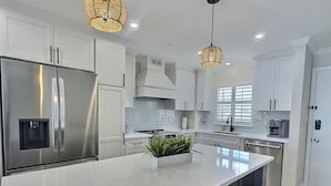 Une cuisine moderne avec des meubles blancs, un réfrigérateur en acier inoxydable, un grand îlot central agrémenté d’une plante, des chaises en osier et des luminaires suspendus.