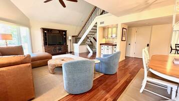A spacious living area with a sofa, two armchairs, a coffee table, and a TV. A staircase leads to a kitchen area.