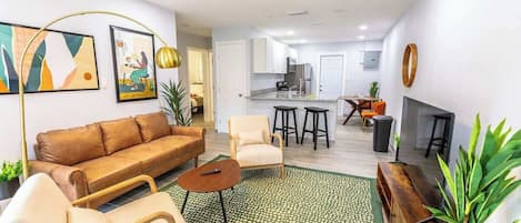 A modern living room with a brown sofa, a beige armchair, a coffee table, and a green patterned rug. There is a kitchen in the background with white cabinets and a dining area with a table and chairs.