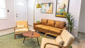 Una sala de estar con un sofá color beige, un sillón de madera y una pequeña mesa de centro. Hay una lámpara de pie, un cuadro abstracto enmarcado y una planta en maceta.
