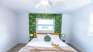 Un dormitorio ordenado con cama, ventana con persianas, ventilador de techo y una pared decorada con follaje verde.